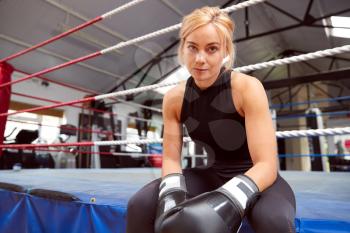 Portrait Of Female Boxer With Gum Shield In Gym Wearing Boxing Gloves Sitting On Boxing Ring