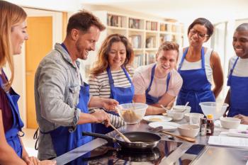Male Teacher Making Pancake On Cooker In Cookery Class As Adult Students Look On