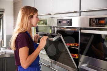 Woman Wearing Apron In Kitchen Checking On Peppers Roasting In Oven Taking Part In Cookery Class