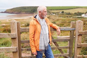 Active Senior Man Walking Along Coastal Path In Fall Or Winter By Gate With Beach And Cliffs Behind