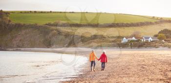 Wide Angle Rear View Of Senior Couple Holding Hands Walking Along Shoreline On Winter Beach Vacation