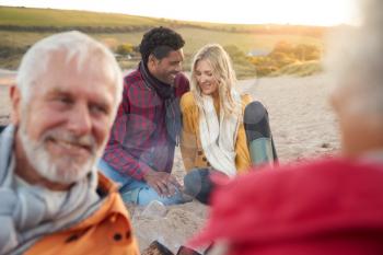 Loving Couple Relaxing By Fire With Senior Parents On Winter Beach Vacation