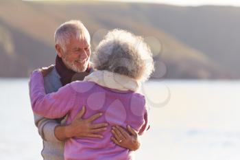 Loving Retired Couple Hugging Standing By Shore On Winter Beach Vacation Against Flaring Sun