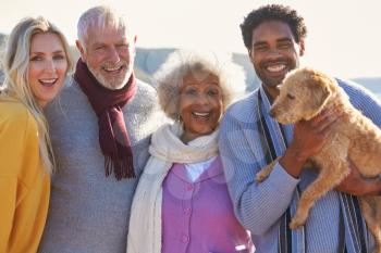 Portrait Of Senior Couple Walking Along Shoreline With Adult Offspring And Dog On Winter Beach Vacation