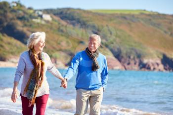 Loving Senior Couple Holding Hands As They Walk Along Shoreline Of Beach By Waves