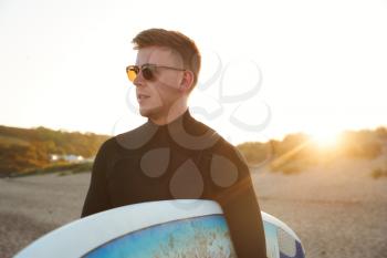 Young Man In Sunglasses Wearing Wetsuit Enjoying Surfing Staycation On Beach As Sun Sets