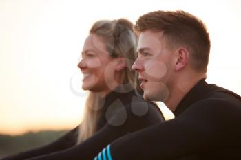Profile View Of Couple Wearing Wetsuits On Surfing Staycation Sitting On Beach Looking Out To  Sea