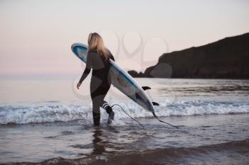 Woman Wearing Wetsuit Carrying Surfboard As She Walks Into Sea