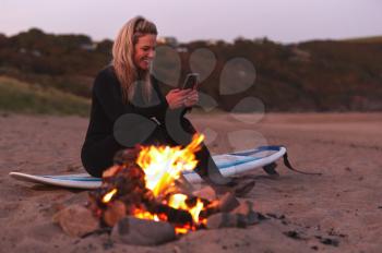 Woman Sitting On Surfboard By Camp Fire On Beach Using Mobile Phone As Sun Sets Behind Her