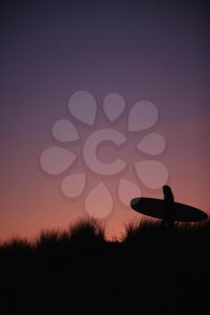 Silhouette Of Female Surfer Carrying Surfboard Across Dunes Against Setting Sun