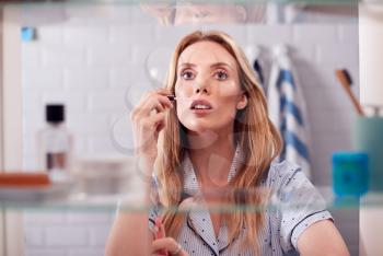 View Through Bathroom Cabinet Of Young Woman Wearing Pajamas Putting On Make Up