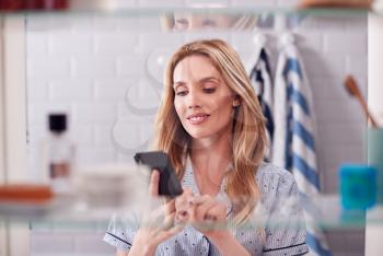 View Through Bathroom Cabinet Of Young Woman Wearing Pajamas Checking Message On Mobile Phone