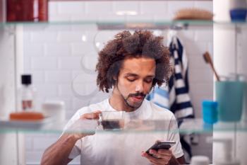 View Through Bathroom Cabinet Of Man Checking Messages On Phone Whilst Drinking Coffee