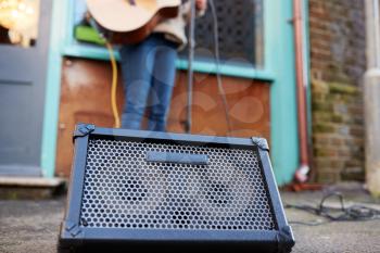 Close Up Of Amplifier As Female Musician Busks Playing Acoustic Guitar Outdoors In Street