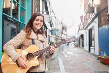 Portrait Of Young Female Musician Busking Playing Acoustic Guitar And Singing Outdoors In Street