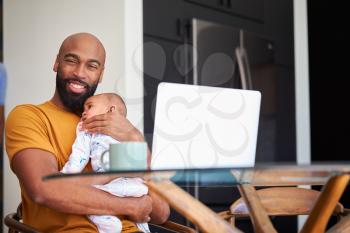 Smiling Stay At Home African American Father Cuddling Baby Daughter Whilst Working On Laptop At Home
