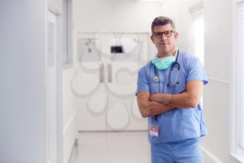 Portrait Of Mature Male Doctor Wearing Scrubs Standing In Hospital Corridor