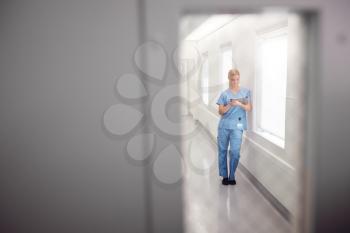 Female Doctor Wearing Scrubs In Hospital Corridor Using Digital Tablet Viewed Through Window In Door