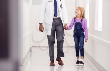 Close Up Of Male Paediatric Doctor Walking Along Hospital Corridor Holding Hands With Girl Patient