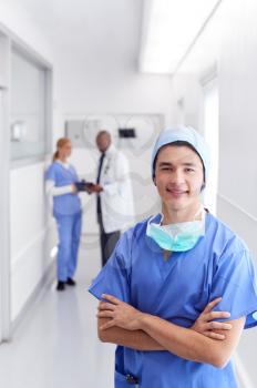 Portrait Of Male Doctor Wearing Scrubs Standing In Busy Hospital Corridor