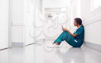 Female Doctor Wearing Scrubs Sitting On Floor In Hospital Corridor Using Mobile Phone