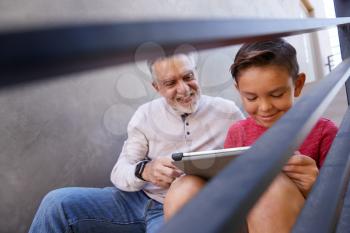 Grandfather Sitting On Steps Outdoors At Home With Grandson Using Digital Tablet