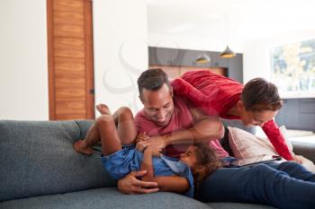 Father Using Digital Tablet At Home Tickling Son And Daughter On Sofa