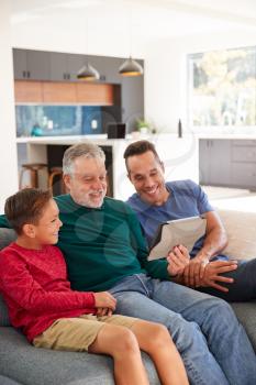 Multi-Generation Male Hispanic Family Sitting On Sofa At Home Watching Movie On Digital Tablet