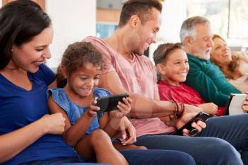 Multi-Generation Family On Sofa Watching TV And Playing With Digital Tablet And Mobile Phones