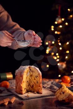 Hand Shaking Icing Sugar Over Christmas Panettone On Table Set For Meal With Tree Lights Behind