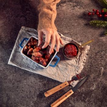Womans Hand Reaches For Dish Of Traditional Christmas Pigs In Blankets On Table Set For Meal