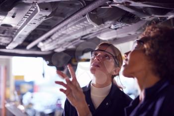 Female Tutor With Student Looking Underneath Car On Hydraulic Ramp On Auto Mechanic Course
