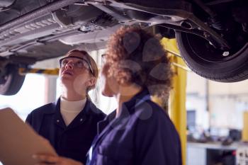 Female Tutor With Student Looking Underneath Car On Hydraulic Ramp On Auto Mechanic Course