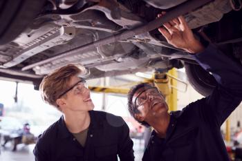 Male Tutor With Student Looking Underneath Car On Hydraulic Ramp On Auto Mechanic Course