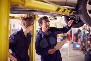 Male Tutor With Student Looking Underneath Car On Hydraulic Ramp On Auto Mechanic Course