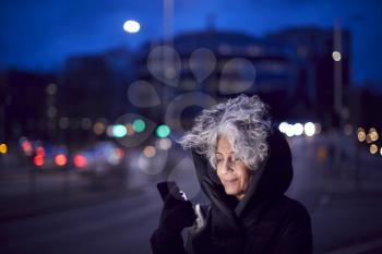 Mature Woman On City Street At Night Ordering Taxi Using Mobile Phone App