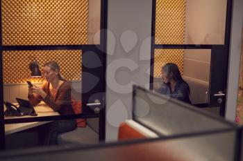 Businesswomen Working Late In Individual Office Cubicles Using Laptop And Digital Tablet