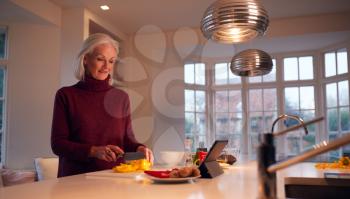Senior Woman In Kitchen At Home Following Recipe On Digital Tablet