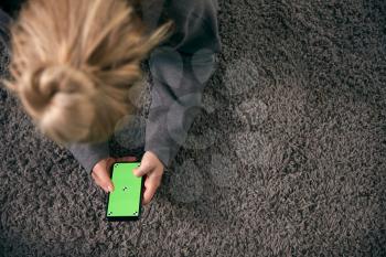 Overhead Shot Of Woman Looking At Green Screen With Copy Space On Mobile Phone Lying On Carpet