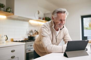 Mature Man In Kitchen Working From Home Using Digital Tablet