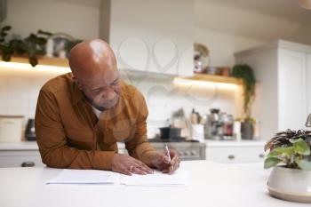 Mature Man Reviewing And Signing Domestic Finances And Investment Paperwork In Kitchen At Home