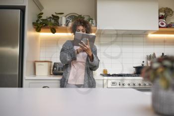 Mature African American Woman In Pyjamas At Home In Kitchen Looking At Digital Tablet