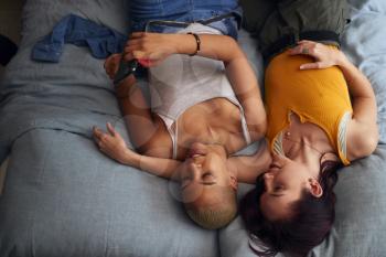 Overhead Shot Of Loving Same Sex Female Couple Lying On Bed At Home Looking At Mobile Phone Together
