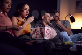 Group Of Friends Eating Popcorn Sitting On Sofa At Home Watching Horror Movie Together