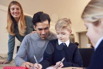 Parents Helping Son And Daughter Wearing School Uniform With Homework At Table In Kitchen