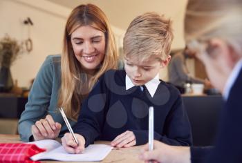 Mother Helping Son And Daughter Wearing School Uniform With Homework At Table In Kitchen