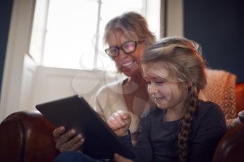 Granddaughter With Grandmother In Chair Looking At Digital Tablet At Home Together