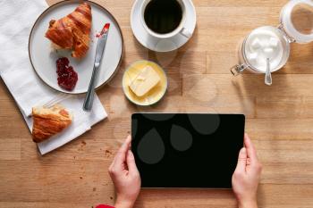 Overhead Flat Lay Of Woman With Digital Tablet On Table Laid For Breakfast With Croissant And Coffee