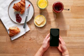 Overhead Flat Lay Of Woman Using Mobile Phone On Table Laid For Breakfast With Croissant And Jam