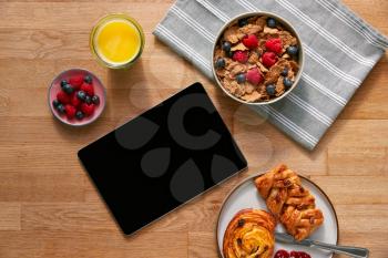 Overhead Flat Lay Of Digital Tablet On Table Laid For Breakfast With Cereal And Pastries